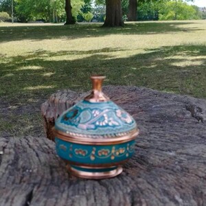 May include: A small, round, lidded container featuring a metallic copper base and rim, with a vibrant turquoise body decorated with intricate, hand-painted floral patterns in gold and copper tones, topped with a matching copper-colored finial knob.