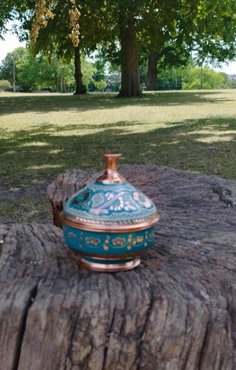 May include: A small, round, lidded container featuring a metallic copper base and rim, with a vibrant turquoise body decorated with intricate, hand-painted floral patterns in gold and copper tones, topped with a matching copper-colored finial knob.