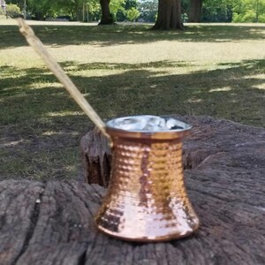May include: A hammered copper coffee pot with a wide base, a tapered body, and a long, slender handle extending diagonally upward. The metallic surface features a textured, dimpled pattern that reflects light across its warm, reddish-brown exterior.