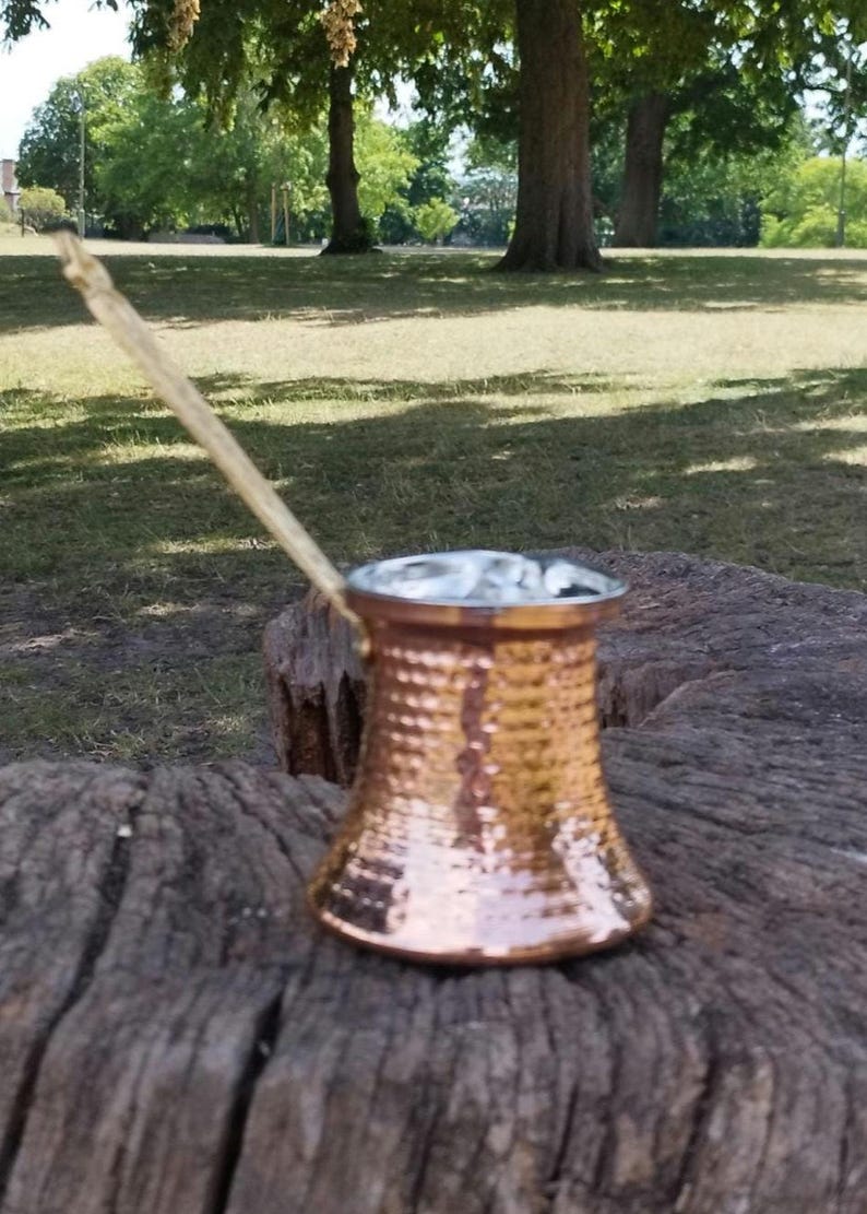May include: A hammered copper coffee pot with a wide base, a tapered body, and a long, slender handle extending diagonally upward. The metallic surface features a textured, dimpled pattern that reflects light across its warm, reddish-brown exterior.