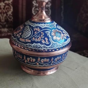 May include: A round copper container with a lid, featuring a deep blue enameled finish decorated with intricate white and copper-toned floral patterns. The base and the top knob of the lid are polished copper, showcasing a metallic sheen.
