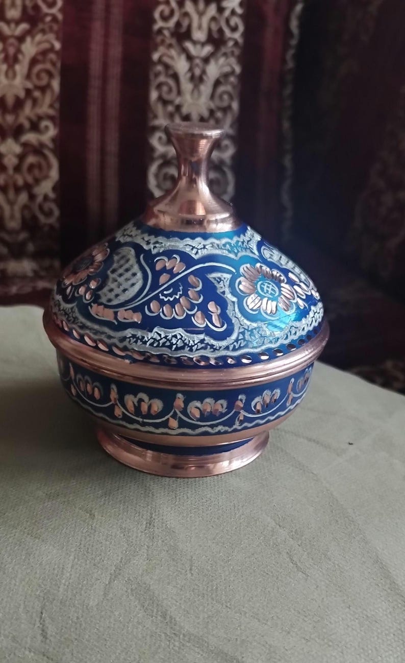 May include: A round copper container with a lid, featuring a deep blue enameled finish decorated with intricate white and copper-toned floral patterns. The base and the top knob of the lid are polished copper, showcasing a metallic sheen.