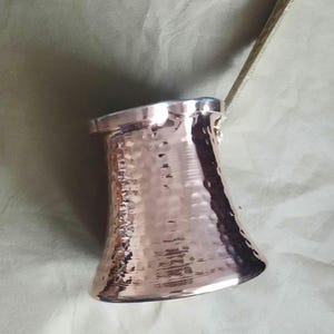 May include: A hammered copper coffee pot with a wide base, a narrow neck, and a flared rim. The pot features a long, slender, metallic handle attached to the side, extending upwards. The surface has a reflective, textured finish with small indentations.