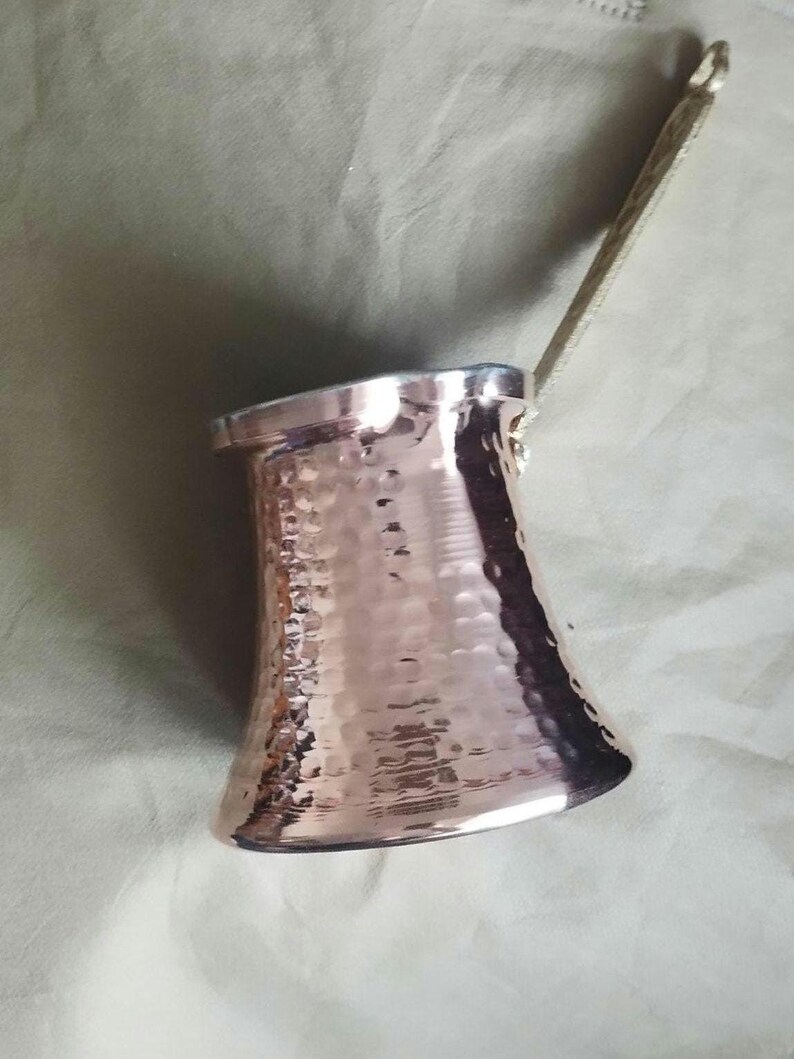 May include: A hammered copper coffee pot with a wide base, a narrow neck, and a flared rim. The pot features a long, slender, metallic handle attached to the side, extending upwards. The surface has a reflective, textured finish with small indentations.