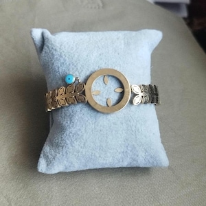 K&ouml;nnte beinhalten: Ein goldfarbenes Metallarmband mit einem zentralen kreisf&ouml;rmigen Anh&auml;nger mit ausgeschnittenem Blattmuster, flankiert von B&auml;ndern mit sich wiederholenden Blattmotiven. Eine kleine, runde, hellblaue Perle ist als Akzent befestigt.