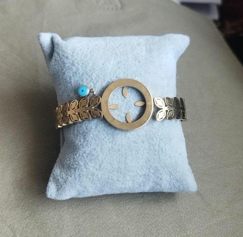 K&ouml;nnte beinhalten: Ein goldfarbenes Metallarmband mit einem zentralen kreisf&ouml;rmigen Anh&auml;nger mit ausgeschnittenem Blattmuster, flankiert von B&auml;ndern mit sich wiederholenden Blattmotiven. Eine kleine, runde, hellblaue Perle ist als Akzent befestigt.