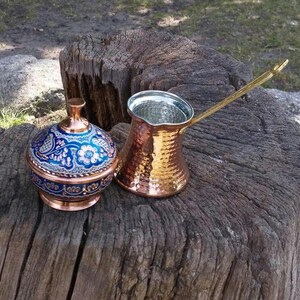May include: A copper coffee pot with a long handle and a hammered texture, paired with a matching lidded container featuring a vibrant blue and white floral pattern. Both items are crafted from polished copper with intricate decorative details.