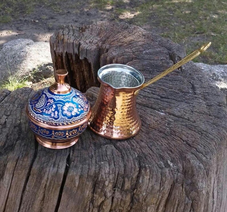 May include: A copper coffee pot with a long handle and a hammered texture, paired with a matching lidded container featuring a vibrant blue and white floral pattern. Both items are crafted from polished copper with intricate decorative details.