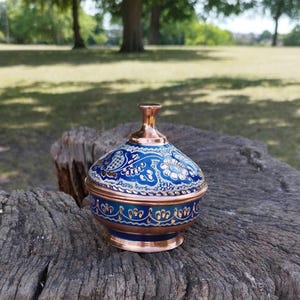 Peut inclure: Un petit bol en cuivre avec un couvercle assorti, dot&eacute; d'une finition en &eacute;mail bleu vif orn&eacute;e de motifs floraux complexes en blanc et or. Le bol pr&eacute;sente une base en cuivre poli et un bouton en cuivre assorti sur le couvercle bomb&eacute;.