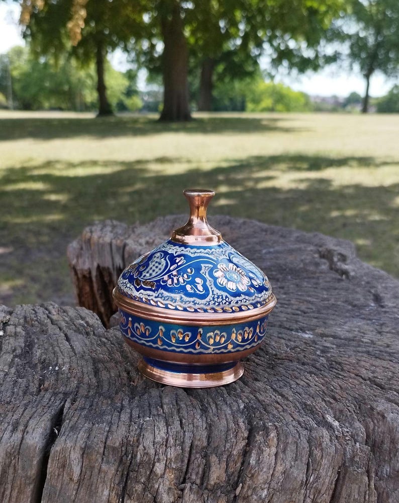 Peut inclure: Un petit bol en cuivre avec un couvercle assorti, dot&eacute; d'une finition en &eacute;mail bleu vif orn&eacute;e de motifs floraux complexes en blanc et or. Le bol pr&eacute;sente une base en cuivre poli et un bouton en cuivre assorti sur le couvercle bomb&eacute;.