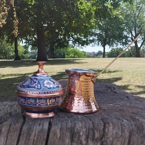 May include: A copper lidded bowl with blue and white floral enamel patterns alongside a hammered copper coffee pot with a long metal handle. Both items feature a metallic finish and are displayed together on a textured wooden surface.