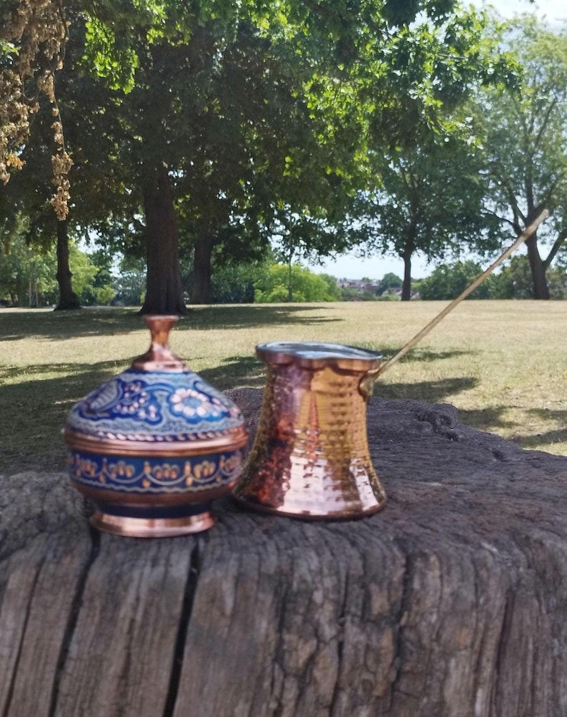 May include: A copper lidded bowl with blue and white floral enamel patterns alongside a hammered copper coffee pot with a long metal handle. Both items feature a metallic finish and are displayed together on a textured wooden surface.