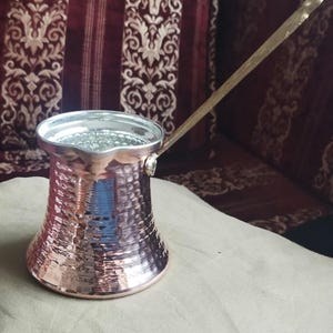 Pode incluir: Uma cafeteira de cobre martelado com base larga, pesco&ccedil;o estreito e borda alargada. O interior &eacute; revestido com um metal de cor prateada. Uma al&ccedil;a met&aacute;lica longa e fina est&aacute; fixada na lateral da cafeteira, estendendo-se diagonalmente para cima.