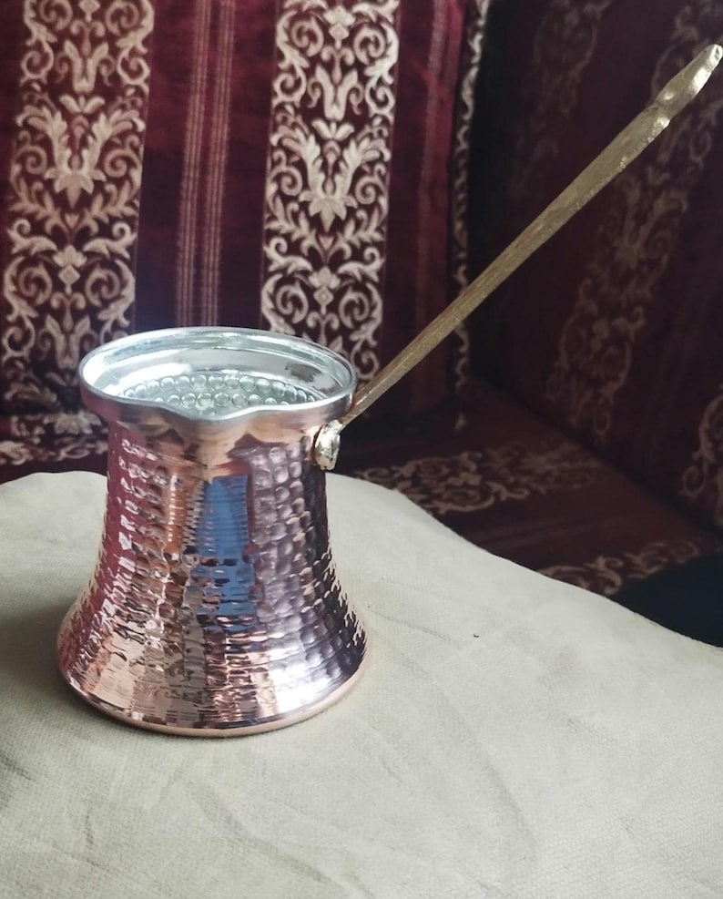 Pode incluir: Uma cafeteira de cobre martelado com base larga, pesco&ccedil;o estreito e borda alargada. O interior &eacute; revestido com um metal de cor prateada. Uma al&ccedil;a met&aacute;lica longa e fina est&aacute; fixada na lateral da cafeteira, estendendo-se diagonalmente para cima.