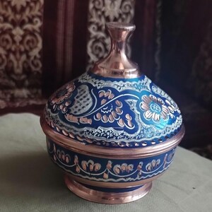 Peut inclure: Un bol rond en cuivre avec un couvercle assorti, dot&eacute; d'une finition en &eacute;mail bleu profond. La surface est orn&eacute;e de motifs floraux et de vignes argent&eacute;s complexes qui r&eacute;v&egrave;lent le m&eacute;tal en cuivre sous-jacent. Le couvercle poss&egrave;de un bouton central.