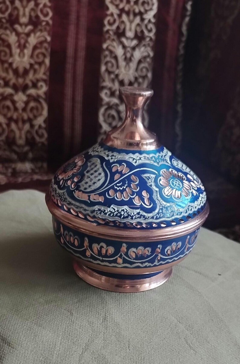 Peut inclure: Un bol rond en cuivre avec un couvercle assorti, dot&eacute; d'une finition en &eacute;mail bleu profond. La surface est orn&eacute;e de motifs floraux et de vignes argent&eacute;s complexes qui r&eacute;v&egrave;lent le m&eacute;tal en cuivre sous-jacent. Le couvercle poss&egrave;de un bouton central.