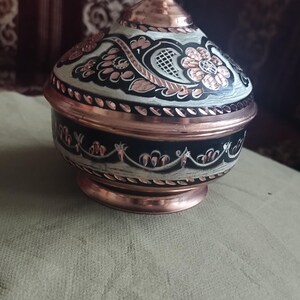 May include: A round copper container with a matching lid, featuring a polished metallic finish and intricate black and white hand-painted floral and leaf patterns. The vessel has a tapered neck, a wide body, and a circular base with decorative trim.