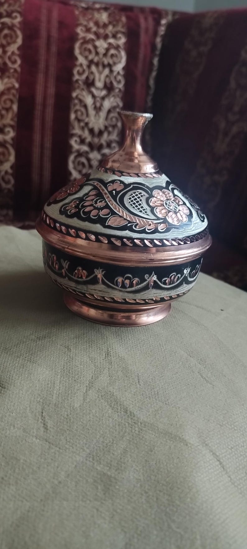May include: A round copper container with a matching lid, featuring a polished metallic finish and intricate black and white hand-painted floral and leaf patterns. The vessel has a tapered neck, a wide body, and a circular base with decorative trim.