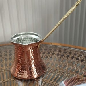 May include: A copper Turkish coffee pot with a hammered exterior finish, a wide pouring spout, and a long, slender brass handle. The interior of the pot is lined with a silver-coloured metal, and the body features a distinct, flared base design.