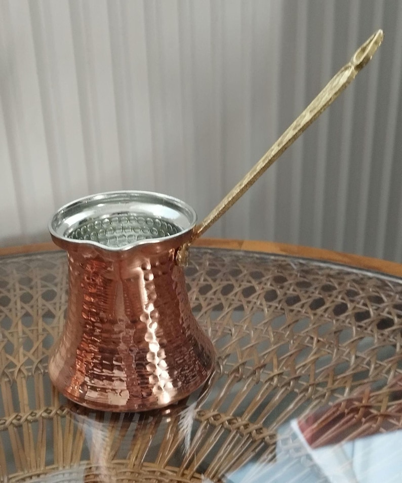 Pode incluir: Uma cafeteira turca de cobre com acabamento exterior martelado, um bico largo e uma pega longa e fina de lat&atilde;o. O interior da cafeteira &eacute; revestido com um metal de cor prateada e o corpo apresenta uma base alargada caracter&iacute;stica.