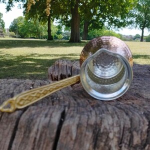 Pode incluir: Uma cafeteira de cobre com exterior martelado e revestimento interior prateado. A cafeteira possui uma pega longa de metal dourado com um padr&atilde;o geom&eacute;trico em relevo ao longo do comprimento e um pequeno la&ccedil;o na extremidade para pendurar.