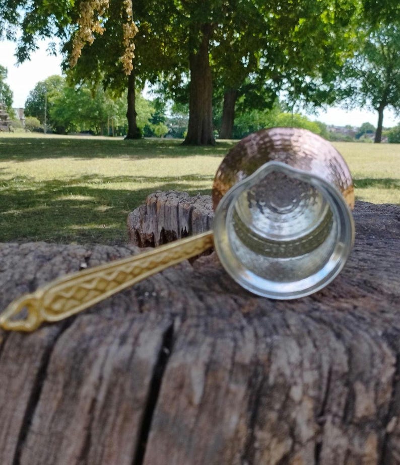Pode incluir: Uma cafeteira de cobre com exterior martelado e revestimento interior prateado. A cafeteira possui uma pega longa de metal dourado com um padr&atilde;o geom&eacute;trico em relevo ao longo do comprimento e um pequeno la&ccedil;o na extremidade para pendurar.