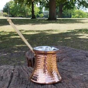 Pode incluir: Uma cafeteira de cor cobre com textura martelada e uma pega longa e fina. A cafeteira apresenta uma base larga e plana que se estreita em dire&ccedil;&atilde;o ao centro antes de alargar ligeiramente na borda superior, que tem um acabamento prateado.