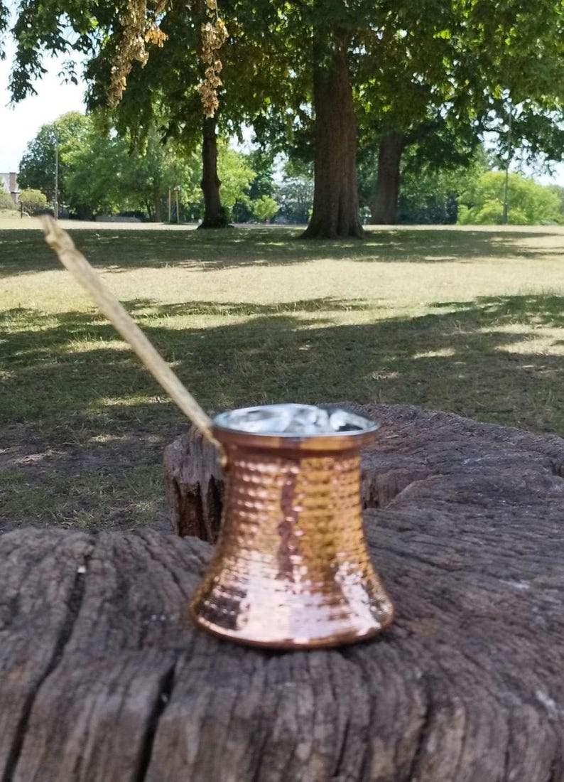 Pode incluir: Uma cafeteira de cor cobre com textura martelada e uma pega longa e fina. A cafeteira apresenta uma base larga e plana que se estreita em dire&ccedil;&atilde;o ao centro antes de alargar ligeiramente na borda superior, que tem um acabamento prateado.