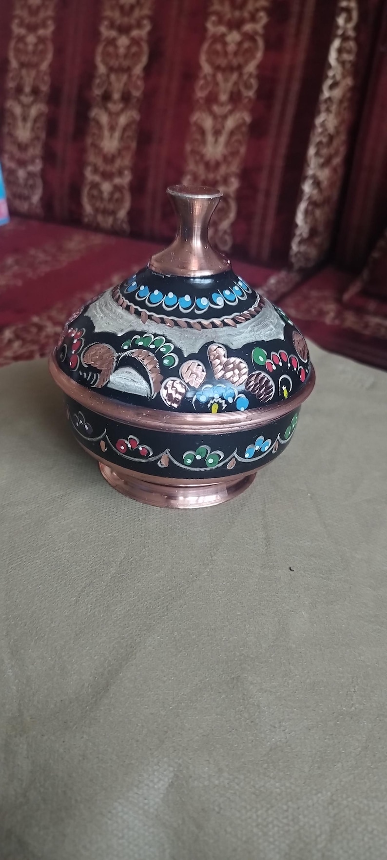 May include: A round metal container with a lid, featuring a black base with hand-painted floral patterns in red, green, and blue. The lid has a copper-colored finial and a matching copper-colored rim at the base, with intricate decorative details.