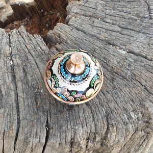 Peut inclure: Un r&eacute;cipient rond avec couvercle, dot&eacute; d'un bord et d'un bouton central de couleur cuivre. La surface pr&eacute;sente un fond noir, des motifs floraux complexes en blanc, rouge, bleu et vert, ainsi qu'une bordure de petits points bleus.