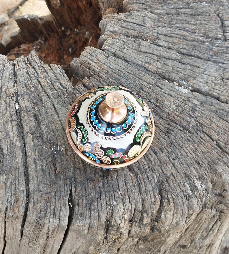 Peut inclure: Un r&eacute;cipient rond avec couvercle, dot&eacute; d'un bord et d'un bouton central de couleur cuivre. La surface pr&eacute;sente un fond noir, des motifs floraux complexes en blanc, rouge, bleu et vert, ainsi qu'une bordure de petits points bleus.