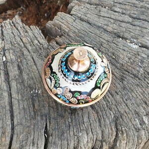 May include: A small, round, lidded container made of copper with a polished finish. The lid features a central knob and is decorated with intricate, hand-painted floral patterns in black, white, blue, green, and red enamel across the surface.
