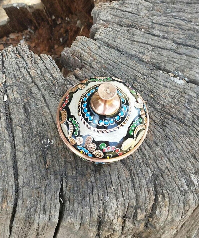 May include: A small, round, lidded container made of copper with a polished finish. The lid features a central knob and is decorated with intricate, hand-painted floral patterns in black, white, blue, green, and red enamel across the surface.