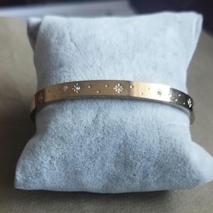 K&ouml;nnte beinhalten: Ein goldfarbener Metallarmreif mit einer Reihe eingravierter Sternmotive und kleinen kreisf&ouml;rmigen Vertiefungen auf der Au&szlig;enfl&auml;che. Das Design enth&auml;lt winzige, klare Steine, die mittig in die Sternformen auf dem Armband eingelassen sind.