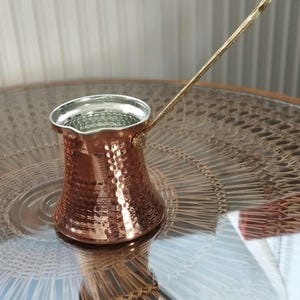 Pode incluir: Uma cafeteira de cobre martelado com uma borda larga e alargada e uma al&ccedil;a longa e fina de lat&atilde;o. O interior da cafeteira &eacute; revestido com um metal de cor prateada e o exterior apresenta uma superf&iacute;cie texturizada que reflete a luz.