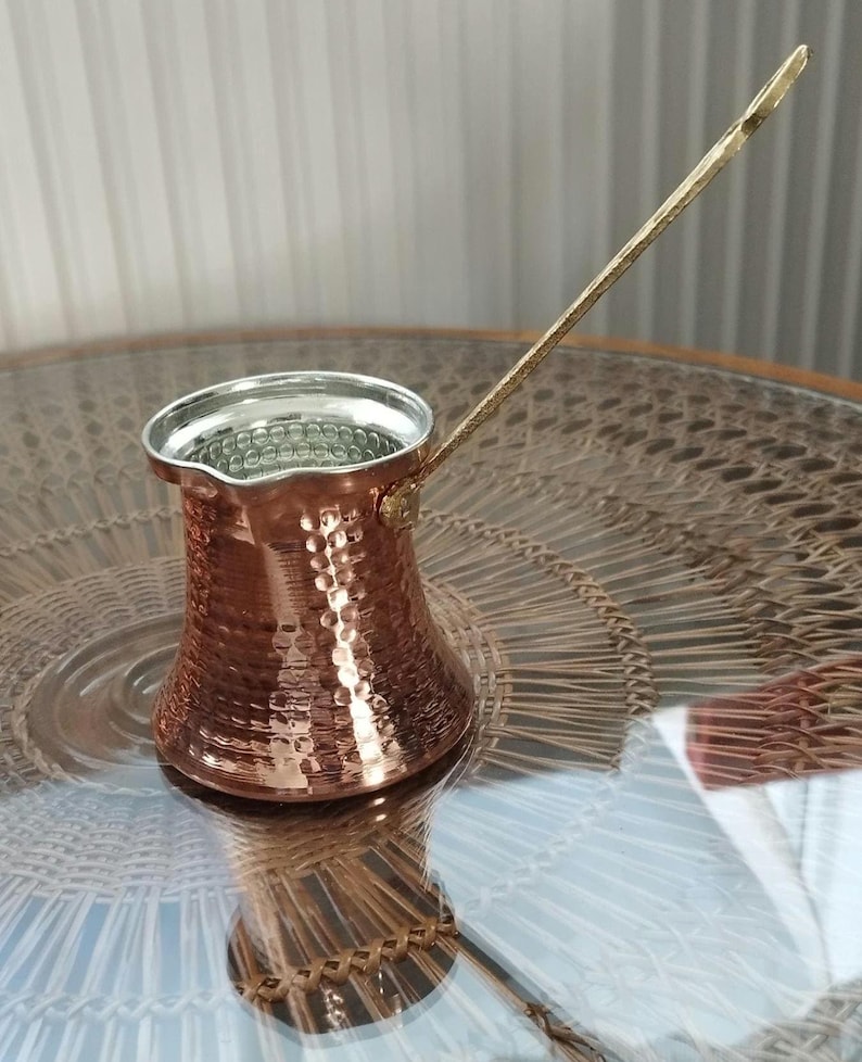 Pode incluir: Uma cafeteira de cobre martelado com uma borda larga e alargada e uma al&ccedil;a longa e fina de lat&atilde;o. O interior da cafeteira &eacute; revestido com um metal de cor prateada e o exterior apresenta uma superf&iacute;cie texturizada que reflete a luz.