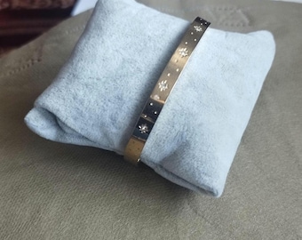 Może przedstawiać: Złota metalowa bransoletka typu bangle z serią małych, grawerowanych motywów w kształcie gwiazdek na zewnętrznej powierzchni. Bransoletka ma gładkie, polerowane wykończenie i jest prezentowana na jasnoszarej, miękkiej kwadratowej poduszce.