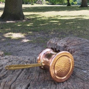 Pode incluir: Uma cafeteira de metal cor de cobre com textura martelada e uma pega longa, plana e de tom dourado. A base da cafeteira apresenta um padr&atilde;o circular em relevo com an&eacute;is conc&ecirc;ntricos, e o corpo tem uma forma cil&iacute;ndrica met&aacute;lica.