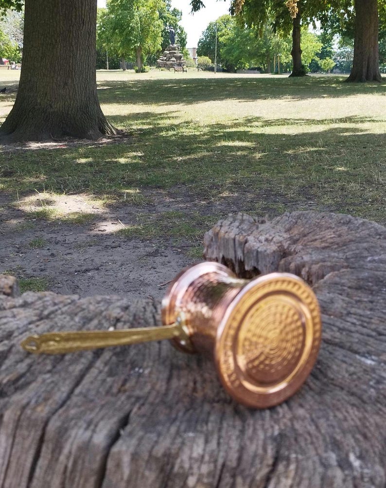 Pode incluir: Uma cafeteira de metal cor de cobre com textura martelada e uma pega longa, plana e de tom dourado. A base da cafeteira apresenta um padr&atilde;o circular em relevo com an&eacute;is conc&ecirc;ntricos, e o corpo tem uma forma cil&iacute;ndrica met&aacute;lica.