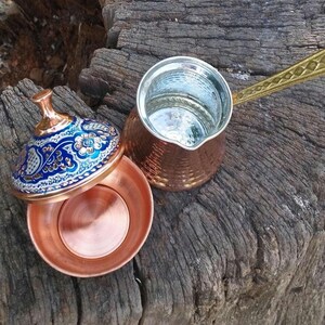 May include: A copper coffee pot with a long, textured brass handle and a matching copper bowl featuring a lid decorated with intricate blue, white, and gold floral patterns. The interior of the pot and bowl are lined with a silver-colored metal.