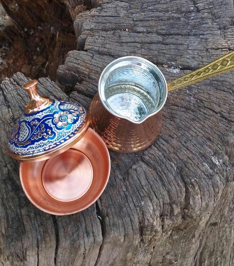 May include: A copper coffee pot with a long, textured brass handle and a matching copper bowl featuring a lid decorated with intricate blue, white, and gold floral patterns. The interior of the pot and bowl are lined with a silver-colored metal.