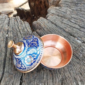 Peut inclure: Un petit bol rond en cuivre avec un couvercle assorti. L'ext&eacute;rieur du couvercle et la base du bol pr&eacute;sentent une finition en &eacute;mail bleu vif d&eacute;cor&eacute;e de motifs floraux complexes blancs et dor&eacute;s. L'int&eacute;rieur du bol est en cuivre poli.