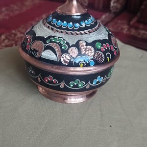 Peut inclure: Un r&eacute;cipient rond en cuivre avec couvercle, dot&eacute; d'une finition en &eacute;mail noir orn&eacute;e de motifs floraux peints &agrave; la main en bleu, vert, rouge et blanc. Le couvercle poss&egrave;de un bouton central en cuivre, la base et le bord sont en cuivre poli.