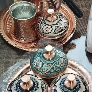 Peut inclure: Un ensemble d'ustensiles de cuisine en cuivre comprenant une th&eacute;i&egrave;re, une tasse en m&eacute;tal martel&eacute; et plusieurs bols avec couvercles. Les bols pr&eacute;sentent des motifs floraux complexes dans des tons vert, blanc et noir avec des accents en cuivre.