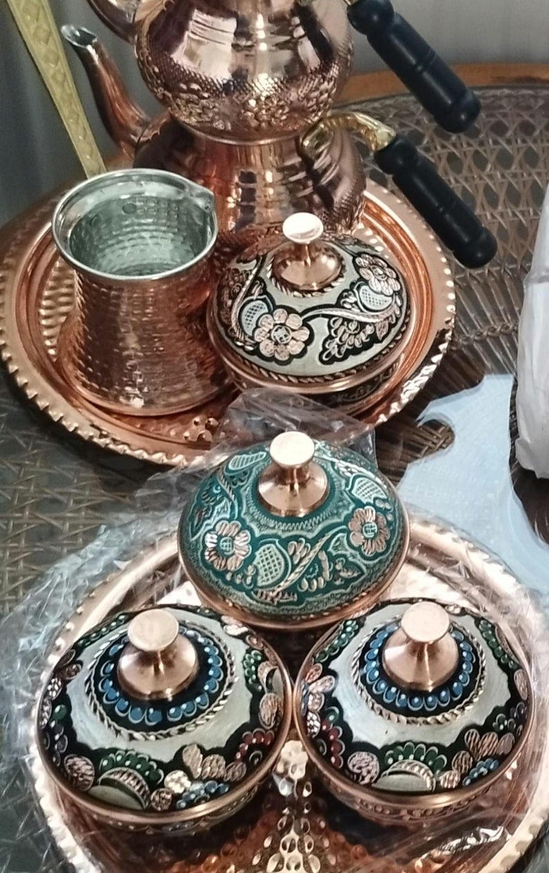 Peut inclure: Un ensemble d'ustensiles de cuisine en cuivre comprenant une th&eacute;i&egrave;re, une tasse en m&eacute;tal martel&eacute; et plusieurs bols avec couvercles. Les bols pr&eacute;sentent des motifs floraux complexes dans des tons vert, blanc et noir avec des accents en cuivre.