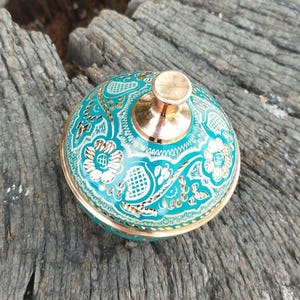 May include: A round metal container with a lid featuring a vibrant turquoise enamel finish and intricate gold-toned floral and geometric engravings. The lid is topped with a polished, smooth, metallic knob in a warm copper-toned finish.