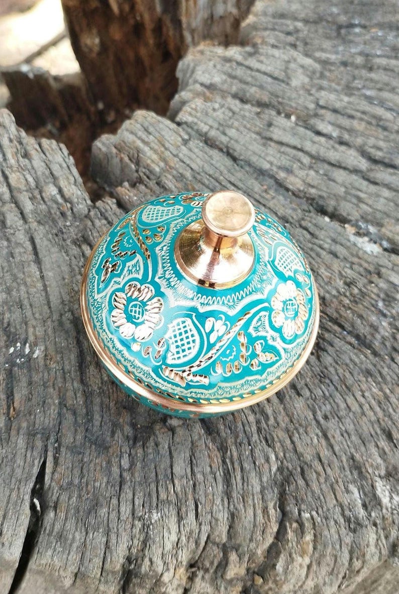 May include: A round metal container with a lid featuring a vibrant turquoise enamel finish and intricate gold-toned floral and geometric engravings. The lid is topped with a polished, smooth, metallic knob in a warm copper-toned finish.