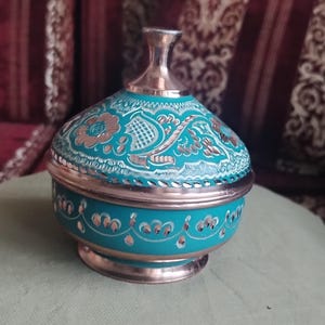 May include: A round copper container with a lid, featuring a vibrant turquoise enamel finish. The surface is decorated with intricate hand-engraved floral and geometric patterns that reveal the metallic copper base beneath the colored coating.