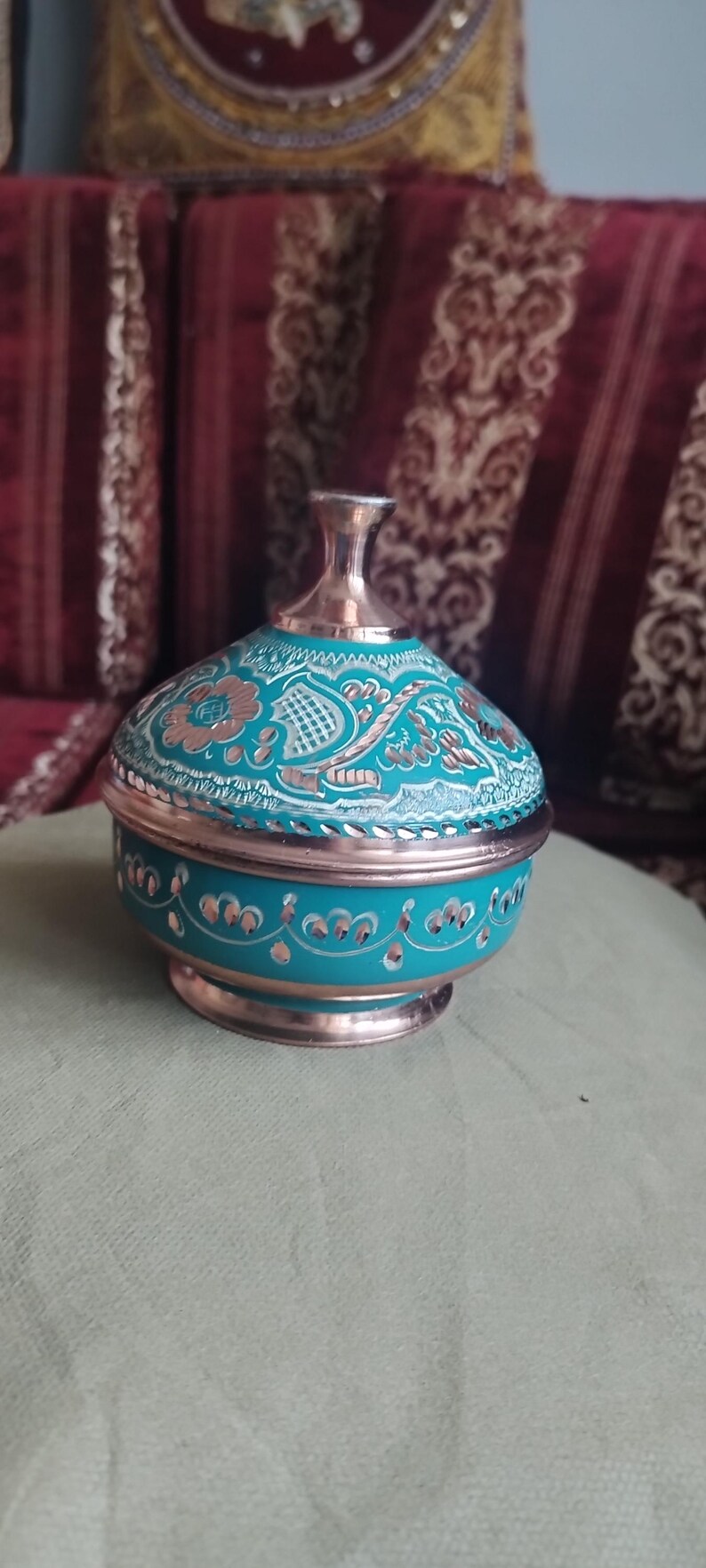 May include: A round copper container with a lid, featuring a vibrant turquoise enamel finish. The surface is decorated with intricate hand-engraved floral and geometric patterns that reveal the metallic copper base beneath the colored coating.
