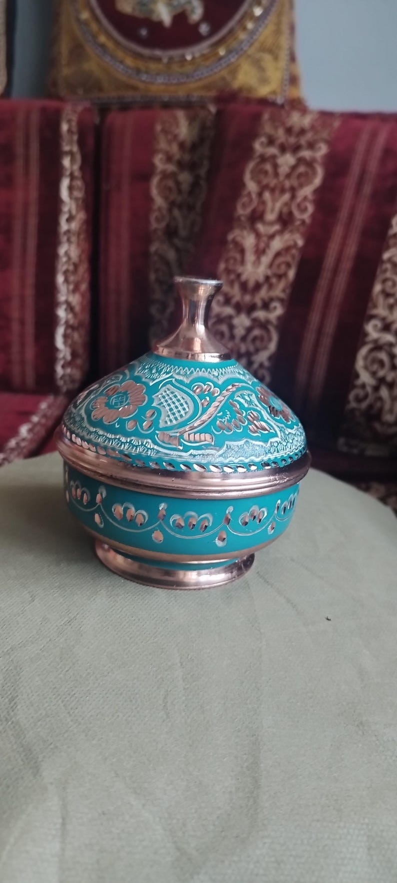 Peut inclure: Un r&eacute;cipient rond en cuivre avec couvercle, dot&eacute; d'une finition &eacute;maill&eacute;e turquoise vive. La surface est orn&eacute;e de motifs floraux et g&eacute;om&eacute;triques grav&eacute;s &agrave; la main, r&eacute;v&eacute;lant la couleur m&eacute;tallique du cuivre sous le rev&ecirc;tement turquoise.